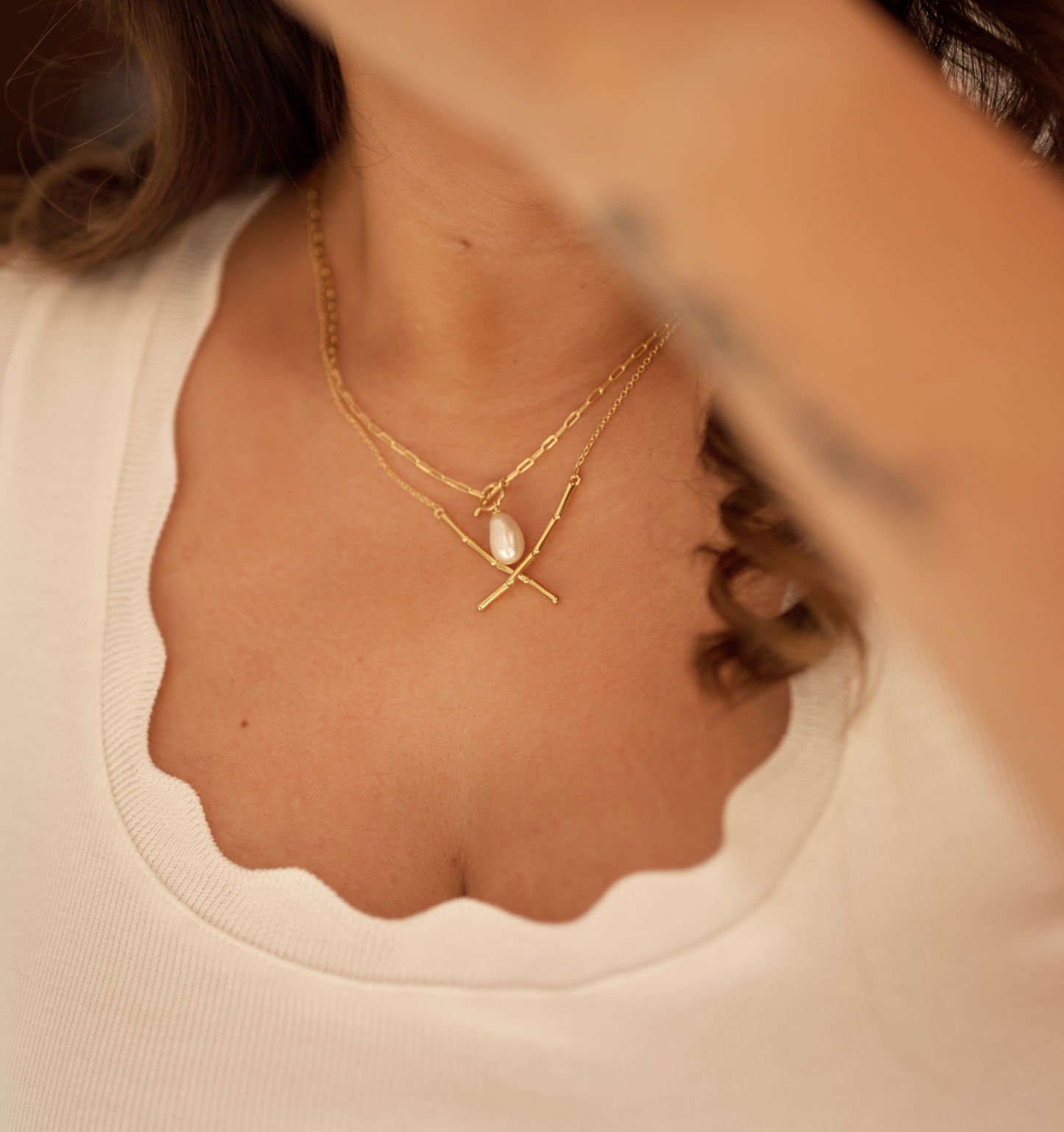 Close-up of a woman wearing a gold Rayi Necklace with a single pearl pendant against a white scalloped neckline top