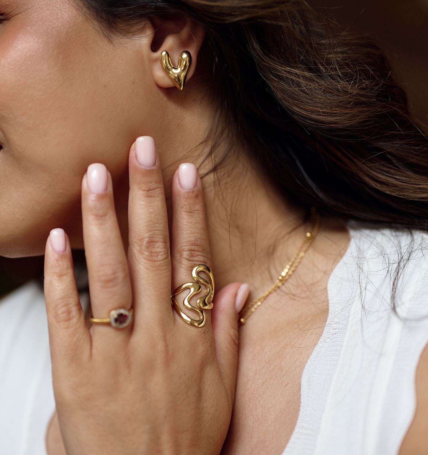 Close-up of hand wearing gold Samvaar Ring with fluid heart design and matching gold earrings