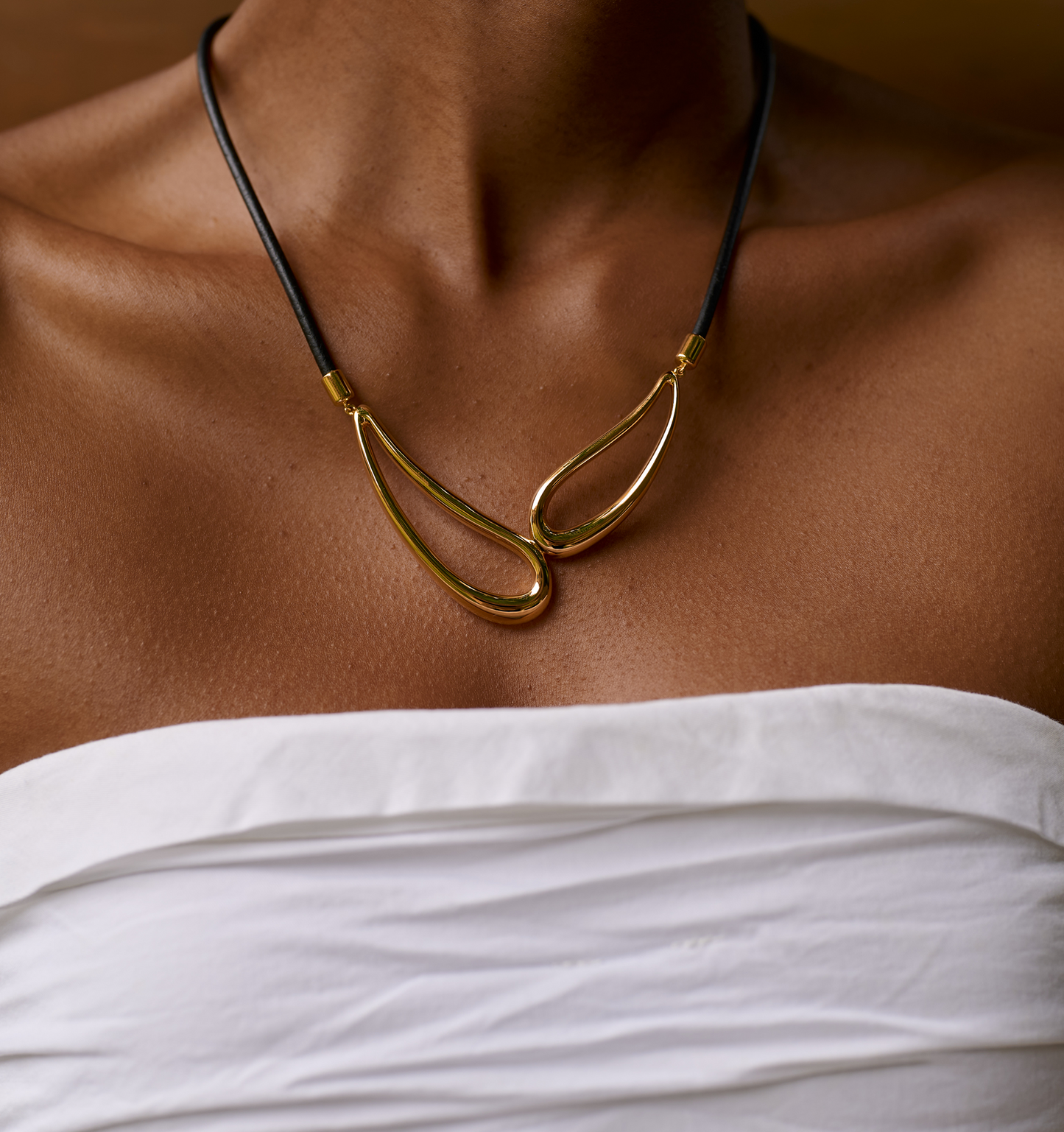Saha Necklace with twin sculpted gold forms on black cord worn with white off-shoulder top