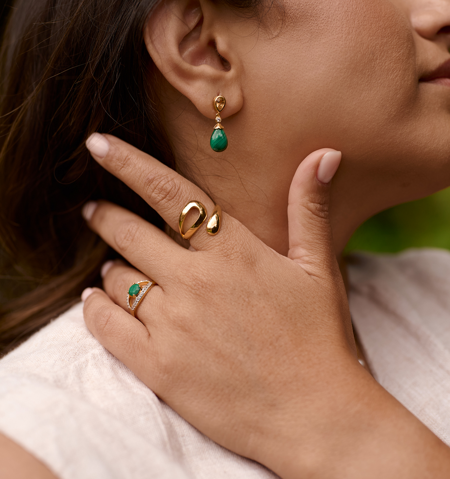 Close-up of hand wearing Oriya Ring with gold earrings featuring a green gemstone on a woman's ear