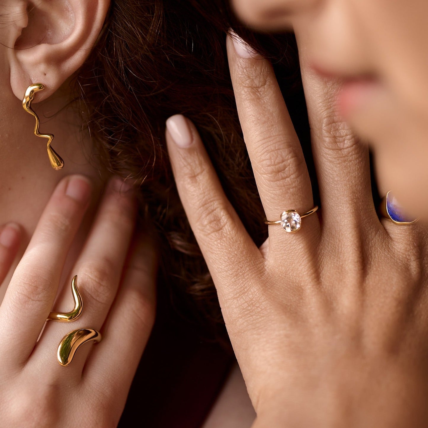 Close-up of Ruhi Ring with white topaz on woman's finger, paired with gold abstract earrings and rings