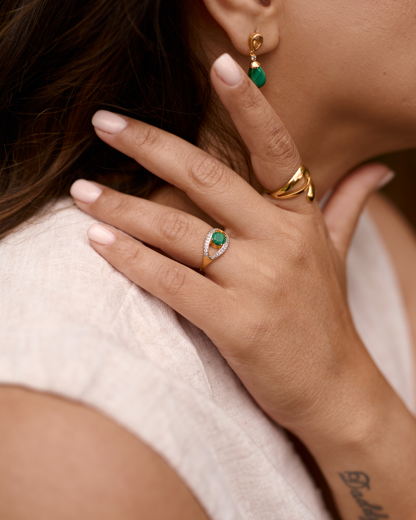 Close-up of hand wearing Saena Ring with green stone and gold jewelry on fingers and ear.