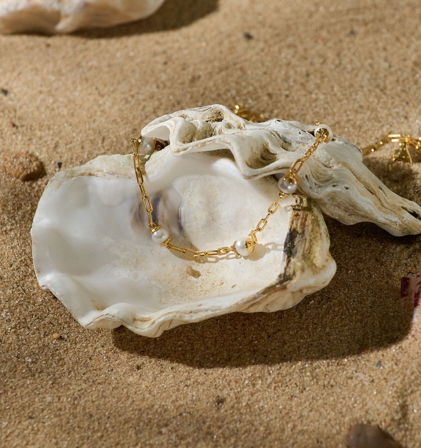 Laya Necklace with gold chain and pearl beads displayed in a seashell on sand background