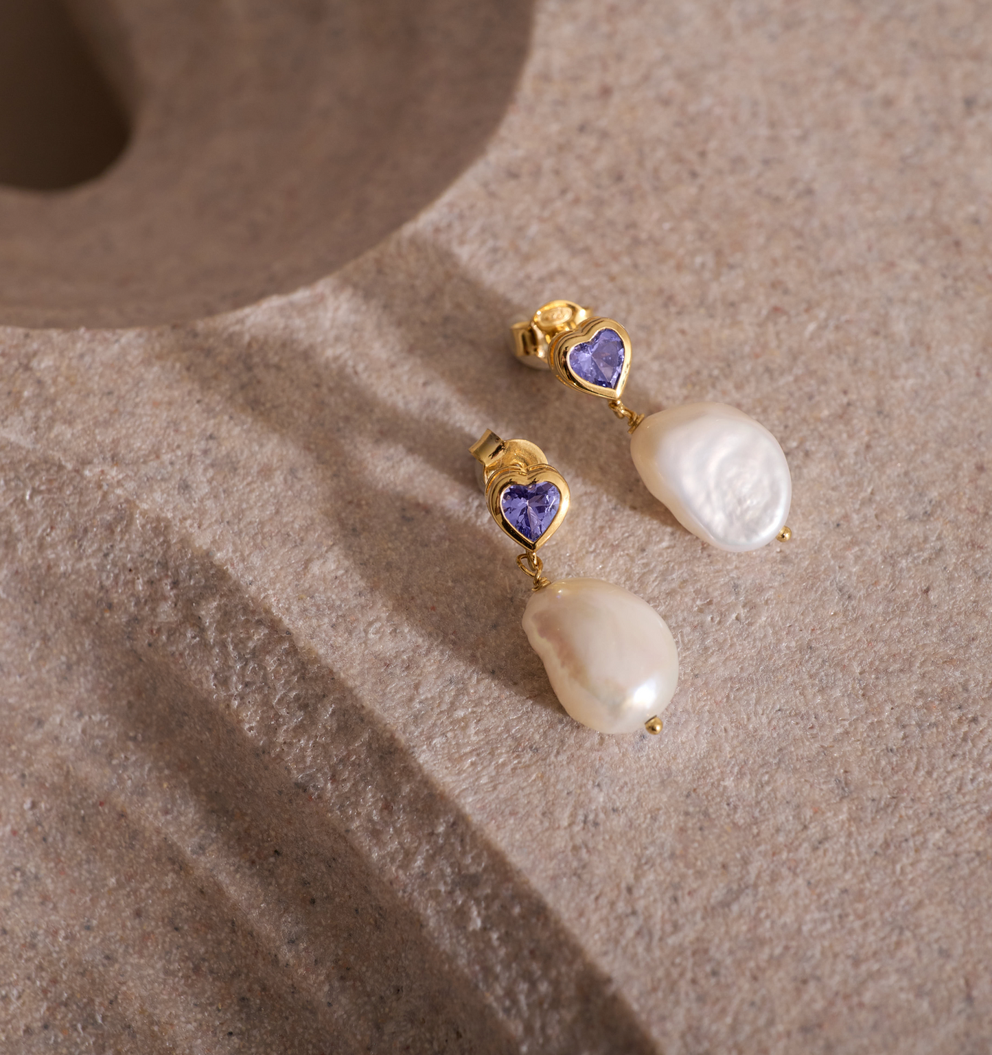 Hridaya Earrings featuring heart-shaped tanzanite and pearl drops on a textured surface