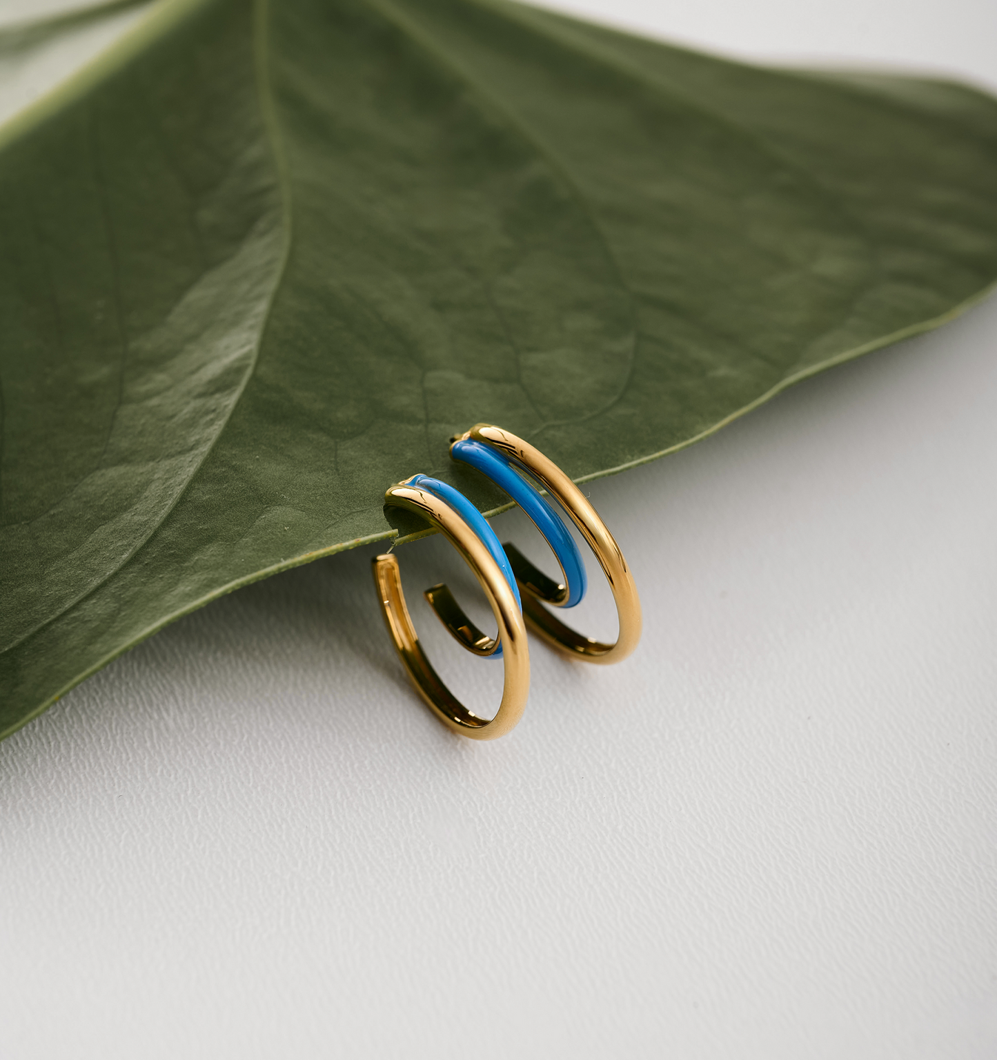 Soma Hoops gold and turquoise enamel crescent-shaped earrings on green leaf background