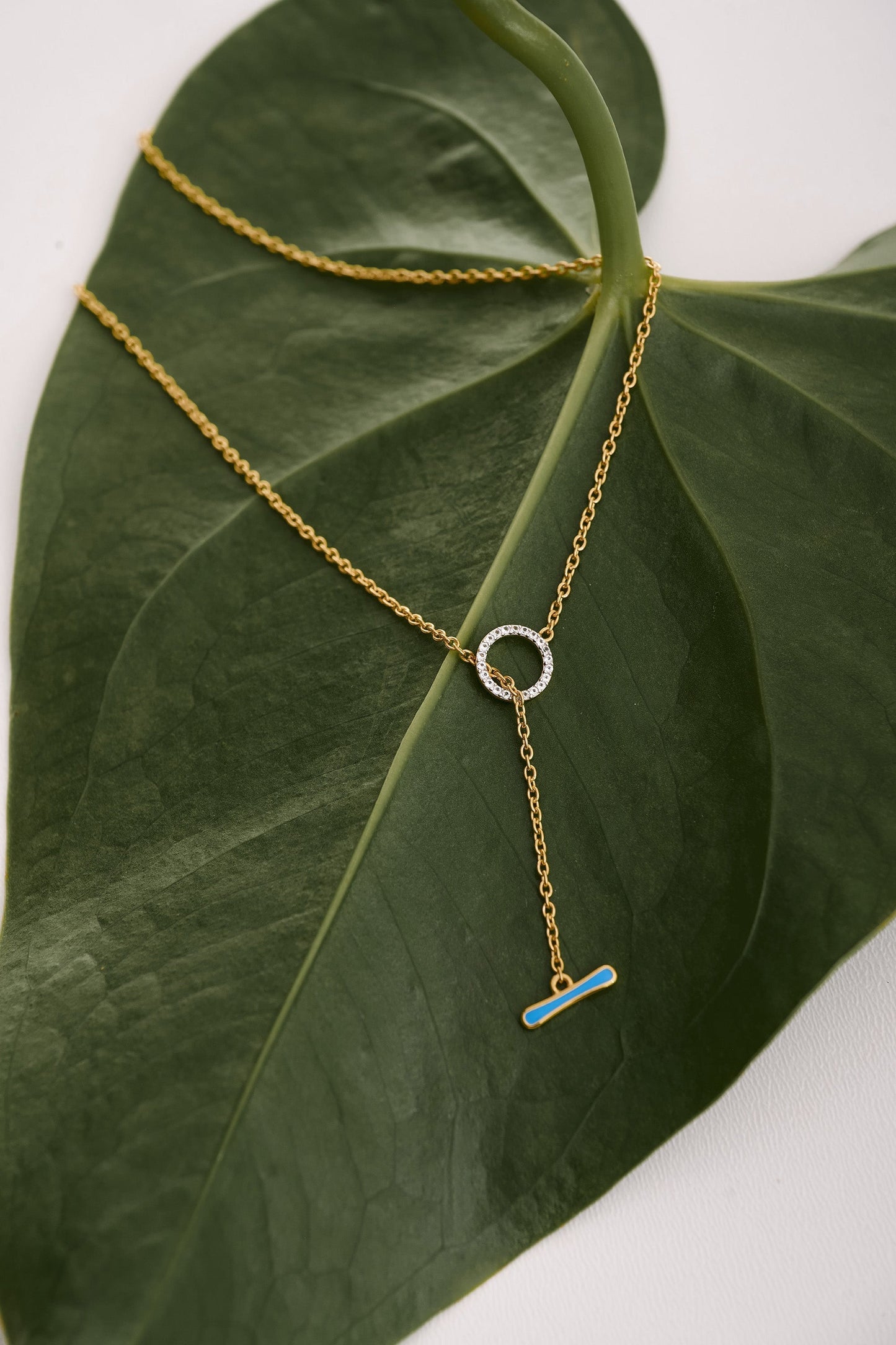Orya Necklace gold chain with white topaz circle and blue bar pendant on green leaf background