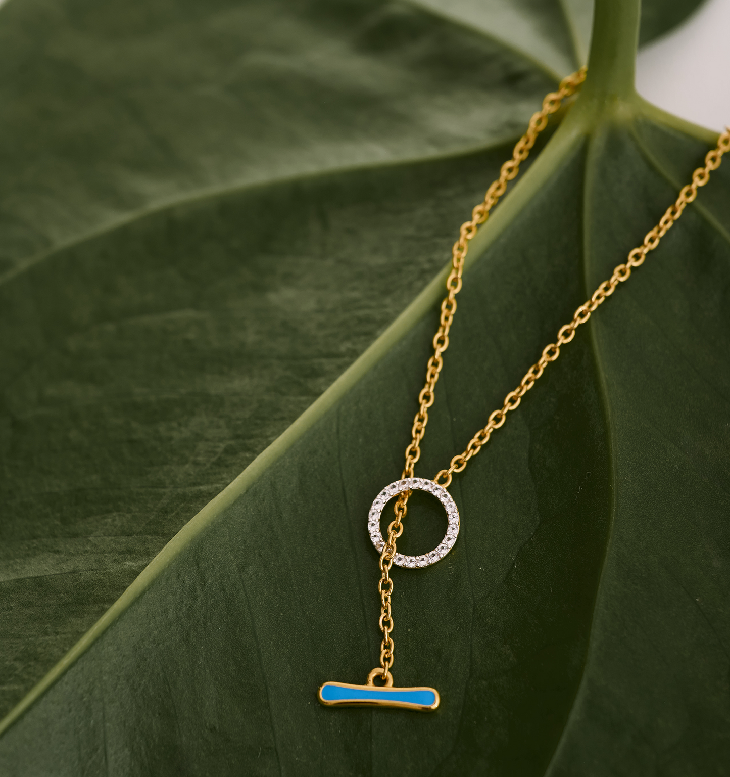 Orya Necklace gold chain with white topaz circle and blue bar pendant on green leaf background