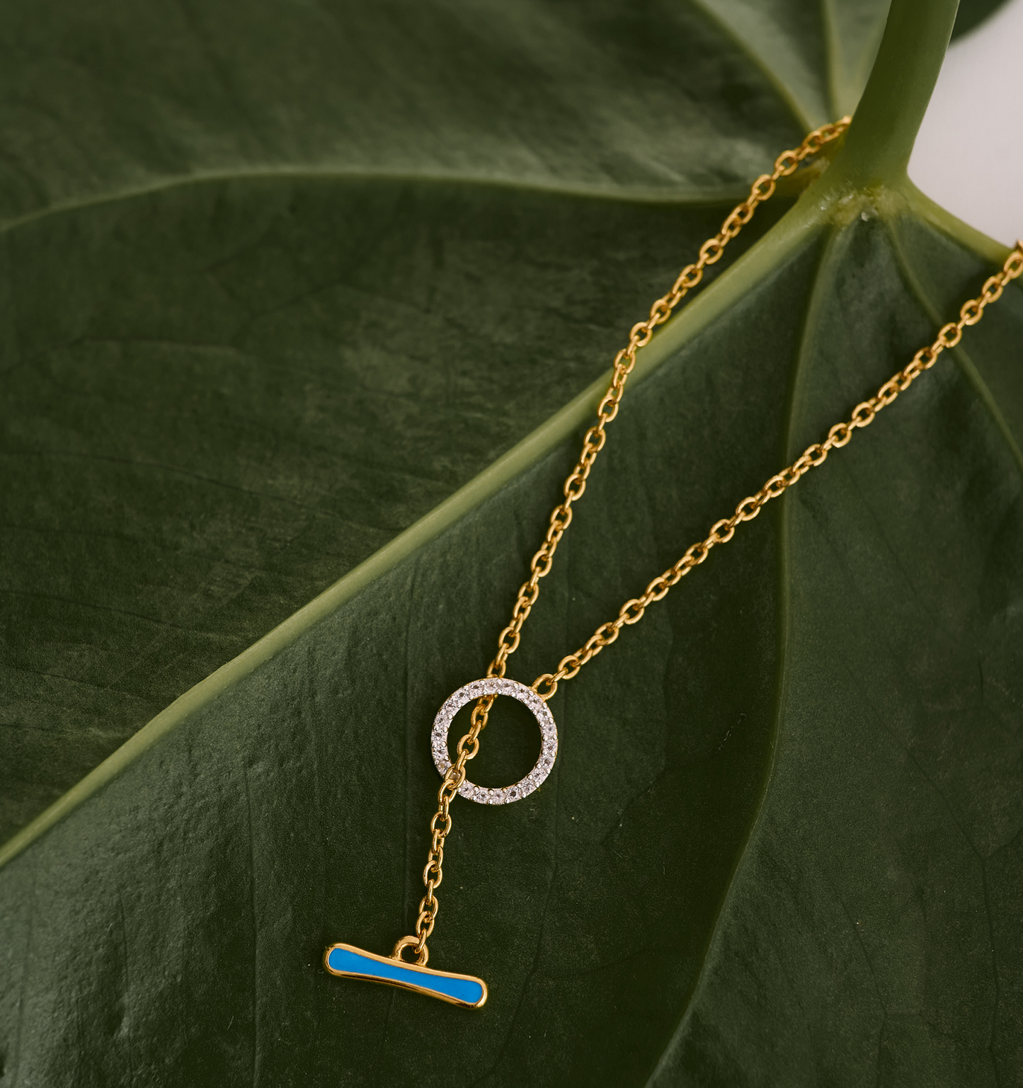 Orya Necklace gold chain with white topaz circle and blue bar pendant on green leaf background
