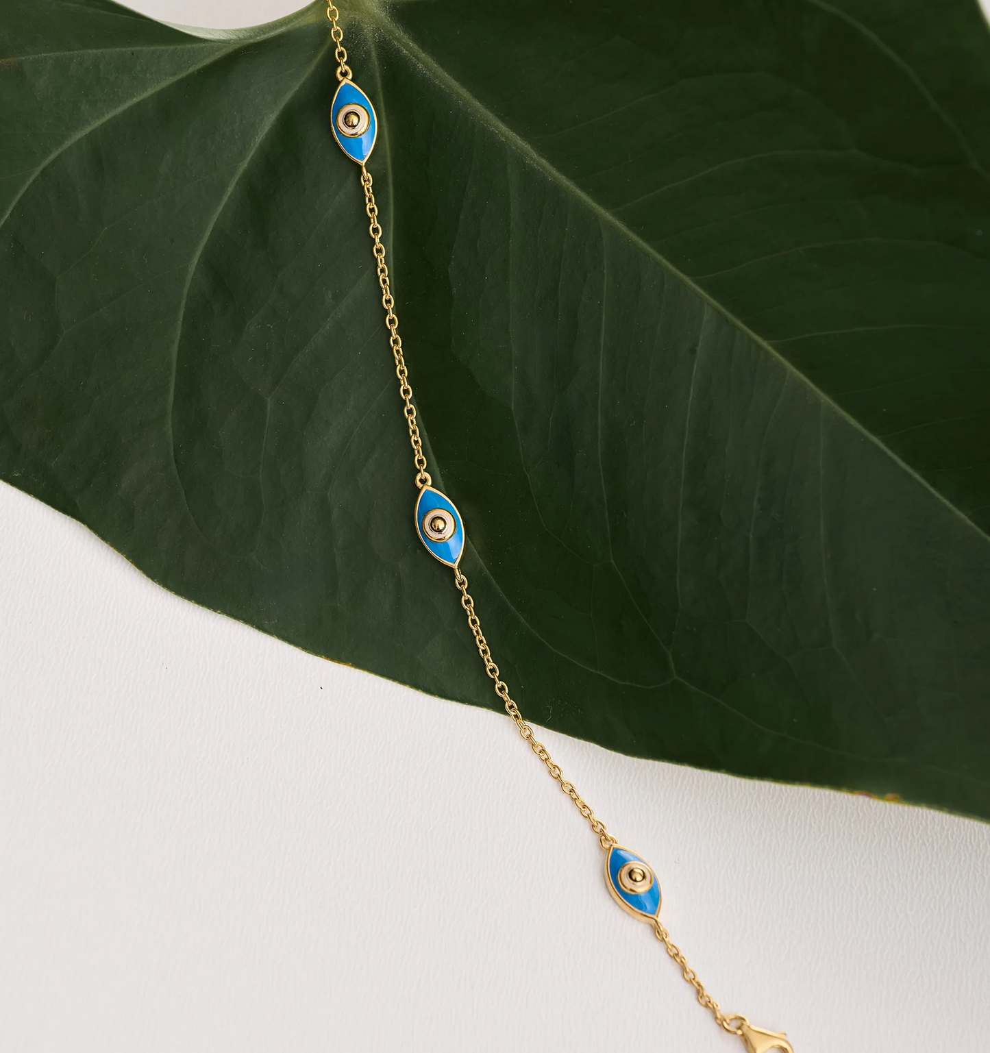 Dhrish Bracelet with turquoise enamel eye charms on gold chain against green leaf background