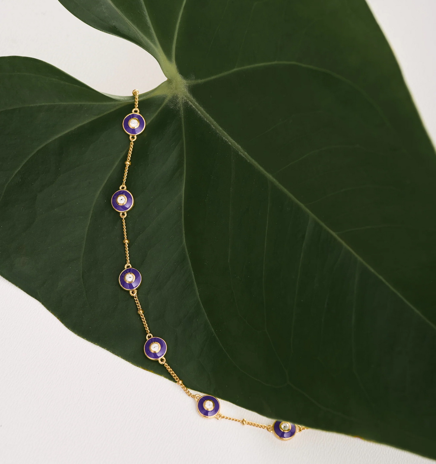 Viraah Bracelet with violet enamel and white topaz on gold chain displayed on green leaf