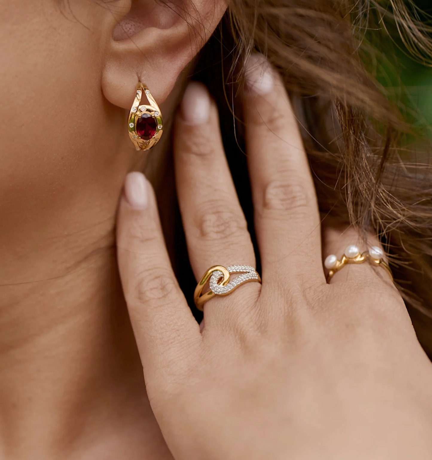 Close-up of ear wearing gold Ruvya Hoops with garnet and hand adorned with gold and pearl rings