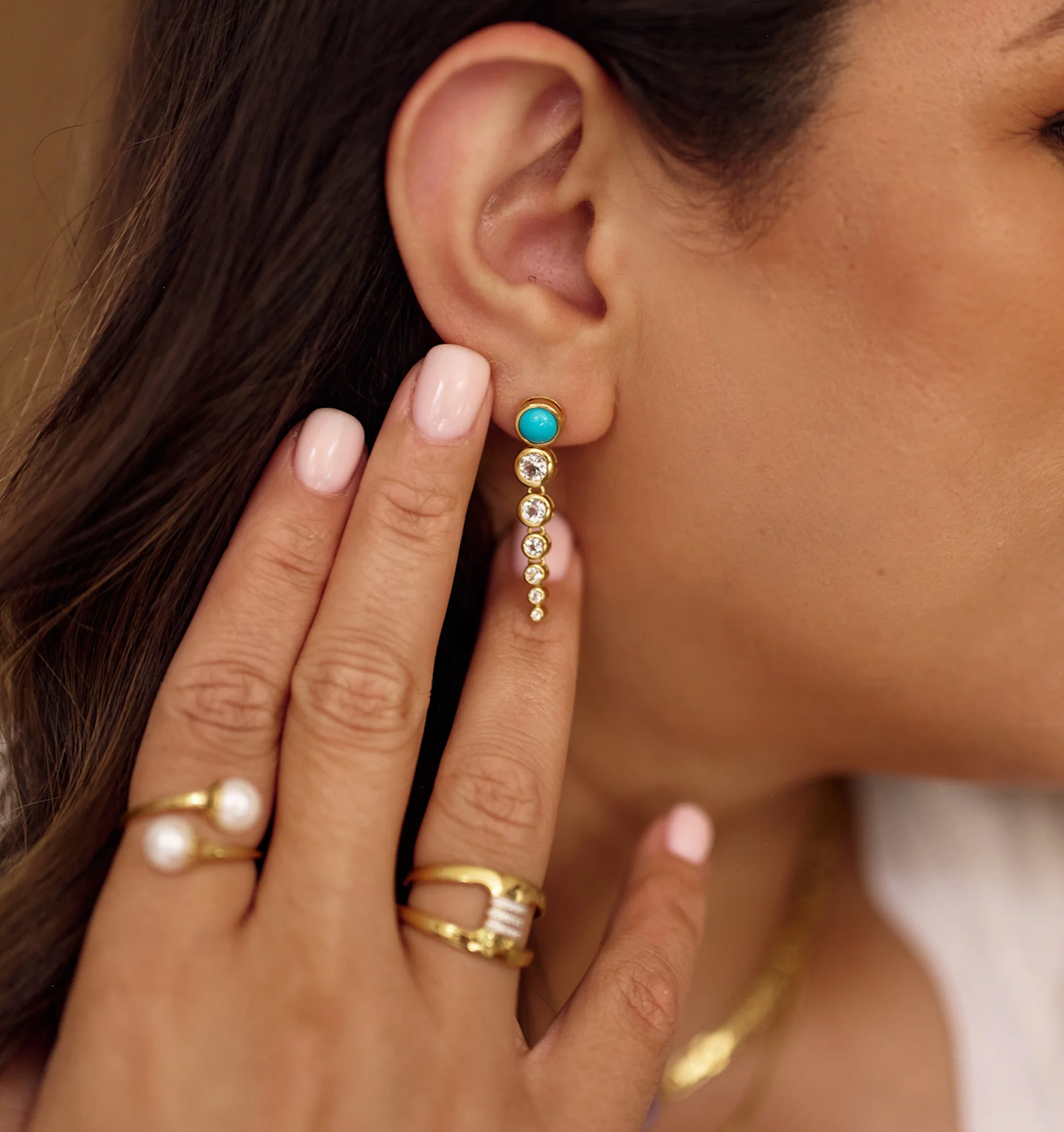 Close-up of Samaara Earrings featuring turquoise and white topaz stones on a woman's ear with gold rings on her fingers