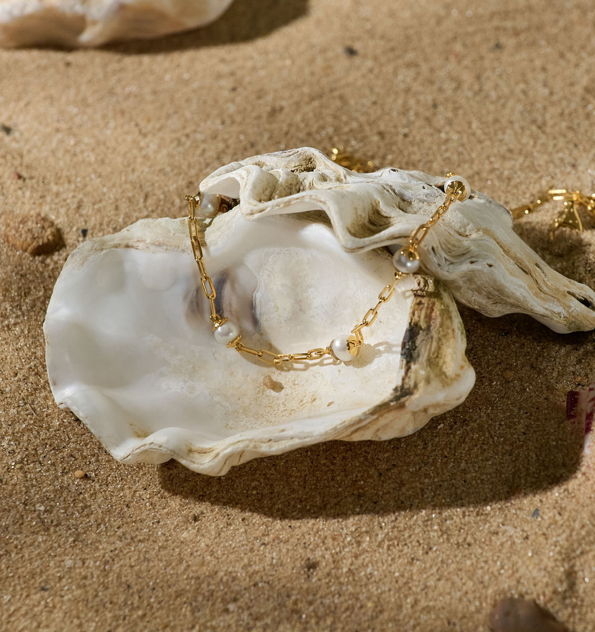 Laya Necklace with gold chain and pearl beads displayed in a seashell on sand background