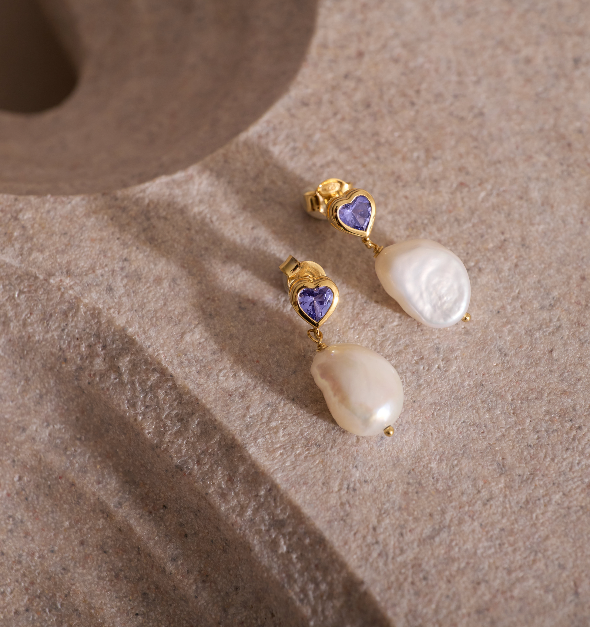 Hridaya Earrings featuring heart-shaped tanzanite and pearl drops on a textured surface