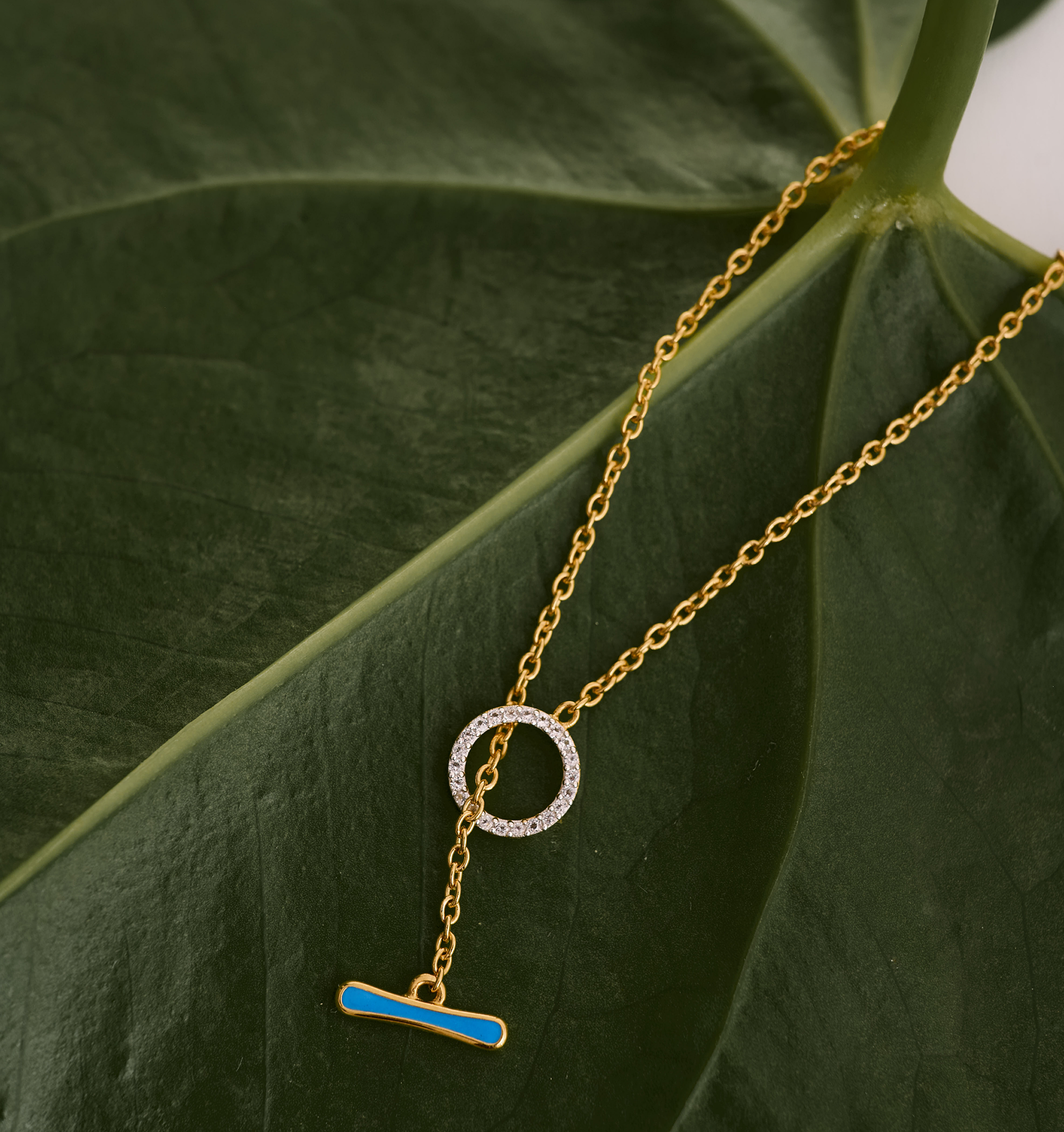 Orya Necklace gold chain with white topaz circle and blue bar pendant on green leaf background