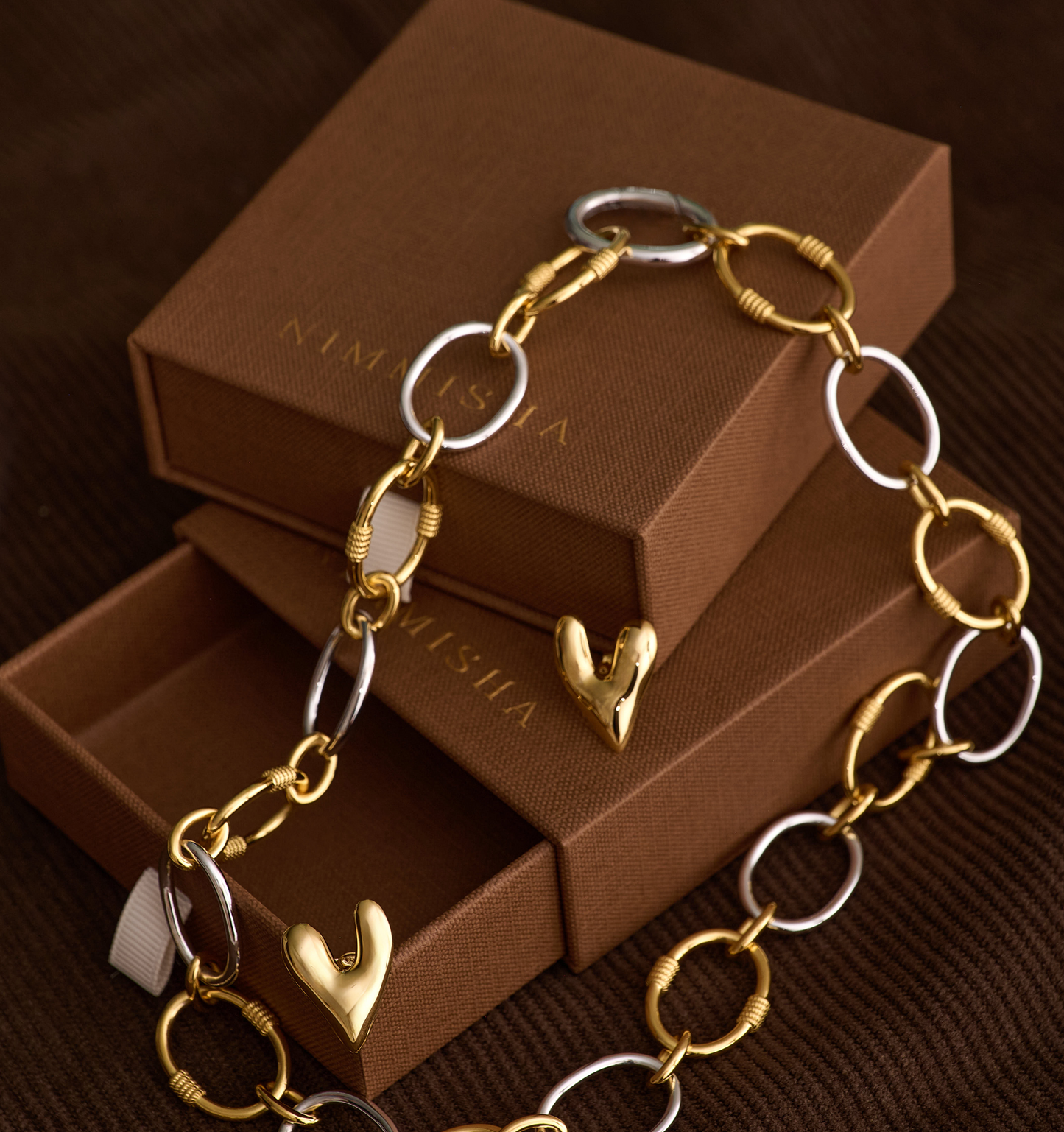 Tasya Necklace featuring alternating gold and silver links with heart-shaped pendants on brown gift box