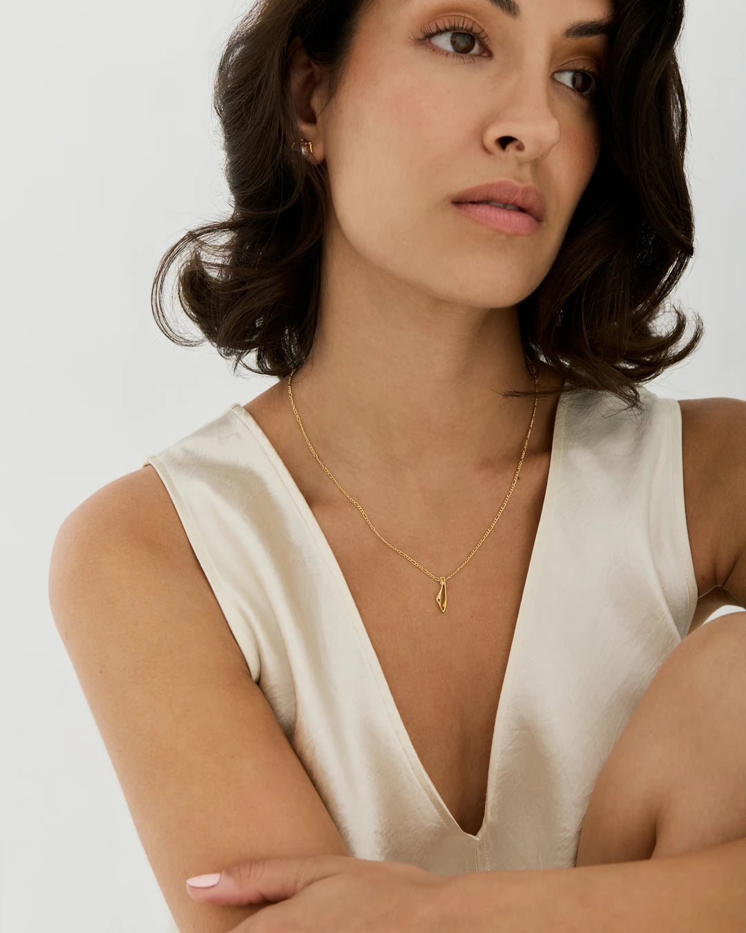 A woman wearing a delicate gold initial necklace with the letter 'I' pendant by NIMMISHA jewellery London, paired with a cream satin V-neck top.