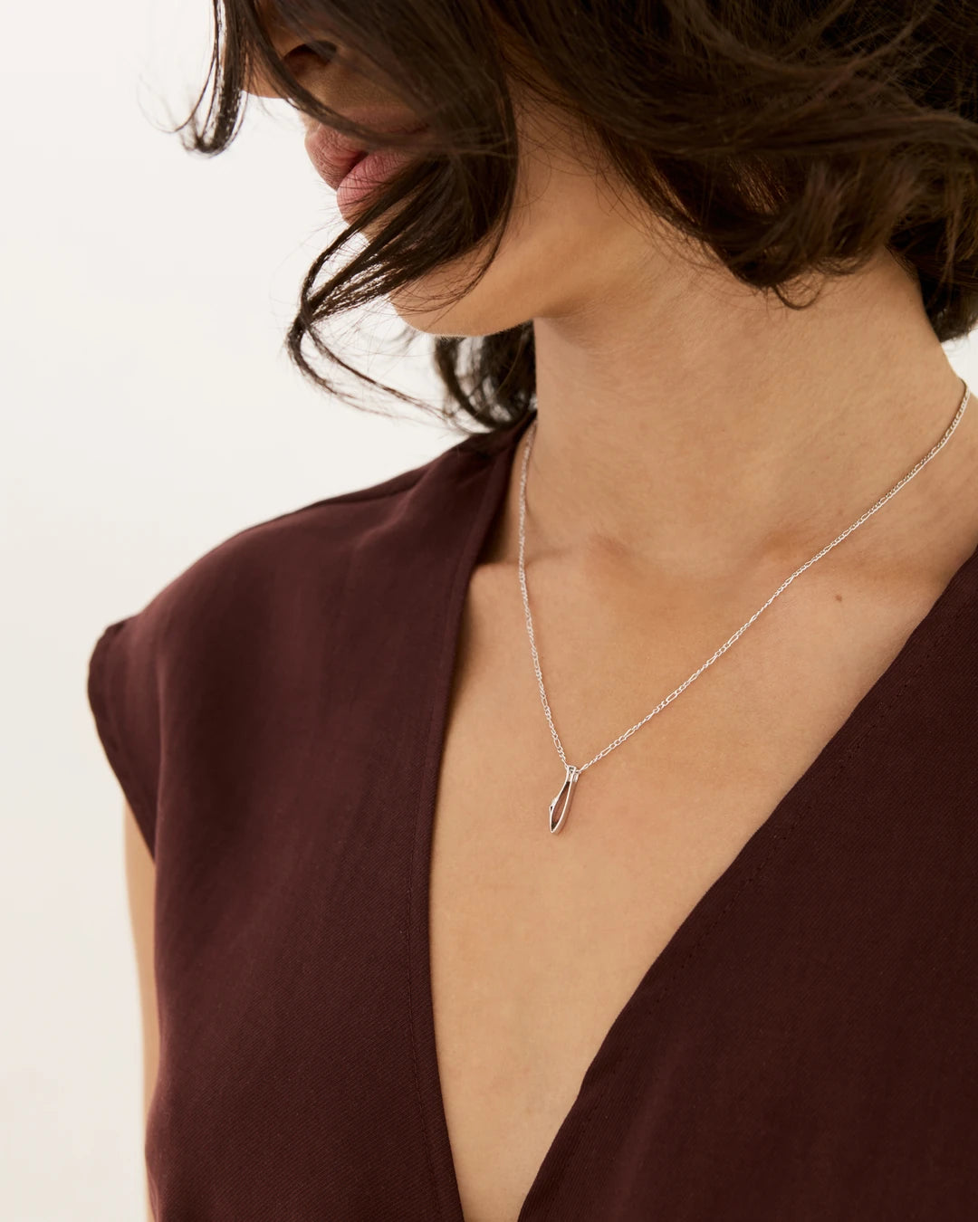 A close-up lifestyle shot of a model wearing a delicate silver initial necklace with the letter 'I' pendant by NIMMISHA jewellery London, paired with a deep burgundy V-neck top.